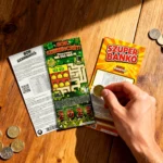 Szuper Bankó, Sok Szerencsét és Kaszinó sorsjegyek egy faasztalon, pénzérmékkel és kockákkal körülvéve, egy kéz épp kaparja az egyik sorsjegyet.